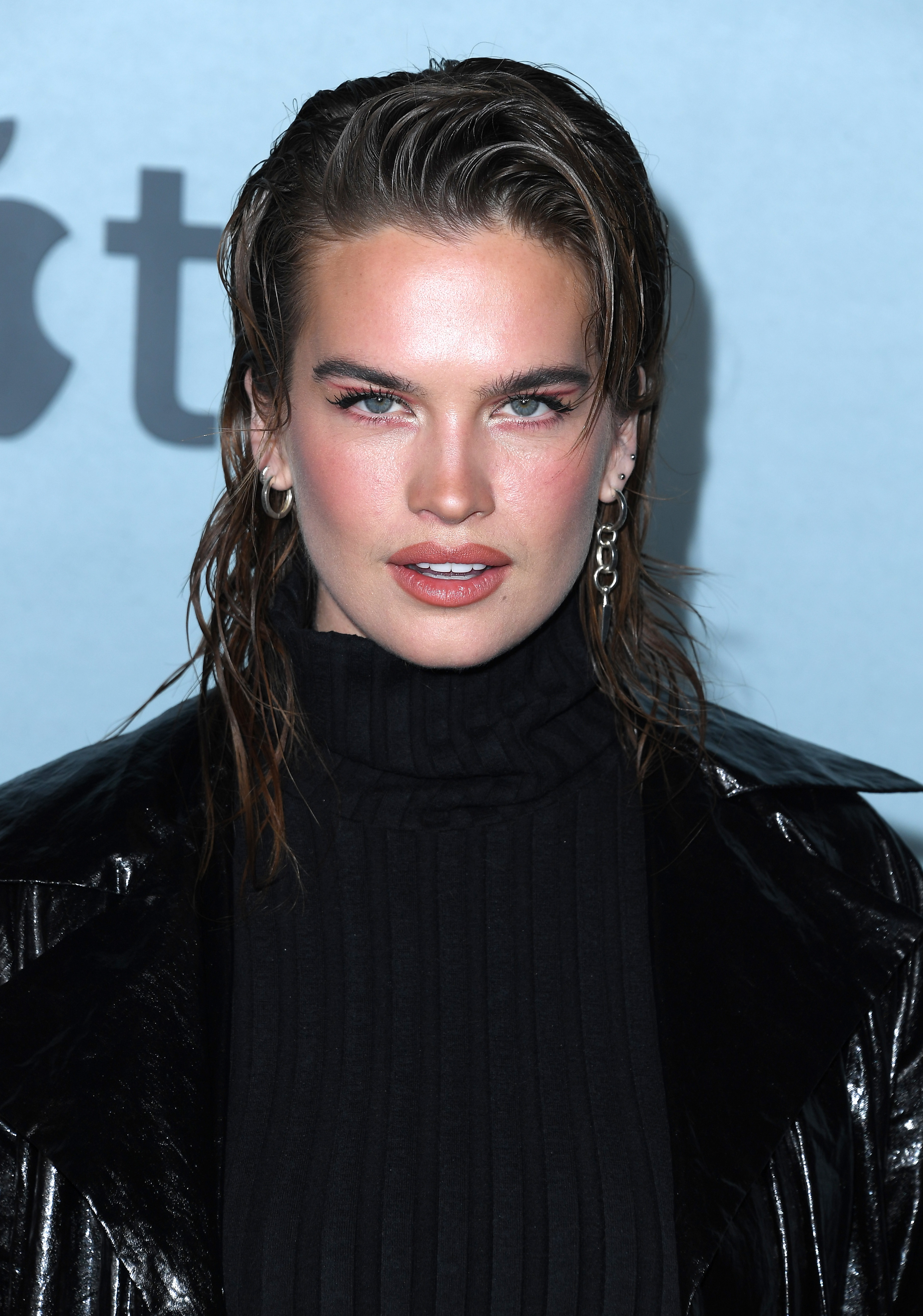 Close-up of a Stormi wearing a turtleneck and shiny jacket, with slicked-back hair and hoop earrings