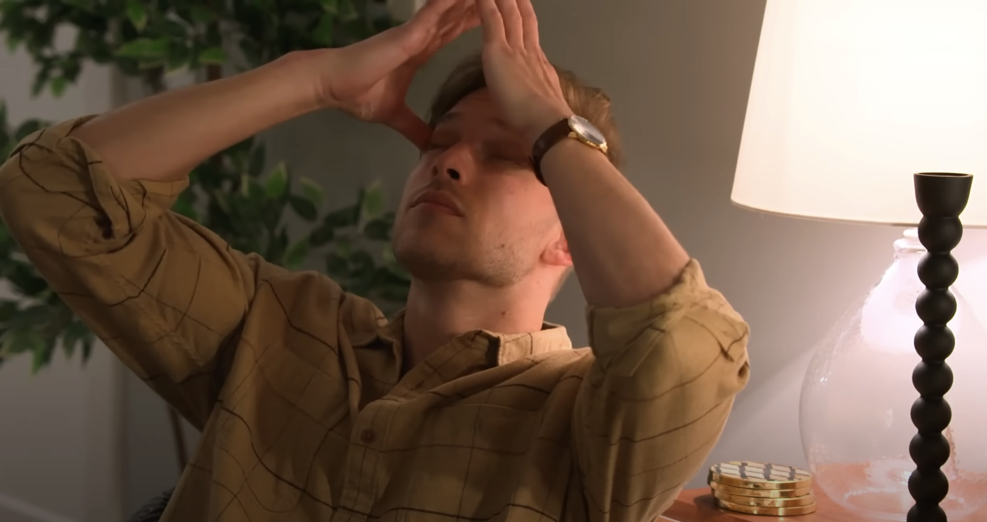 Man in checkered shirt with hands on head, eyes closed, expressing frustration or fatigue, indoors beside a lamp