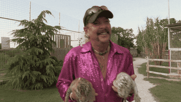 Joe Exotic in a sparkly shirt holding two tiger cubs at an outdoor enclosure