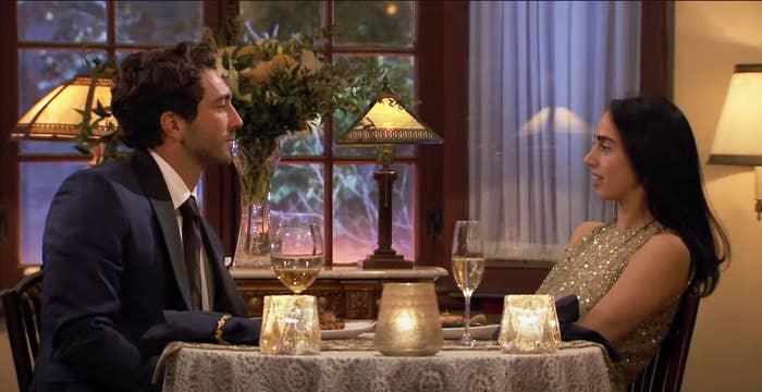 Two people in formal attire having a conversation at a dinner table with wine glasses