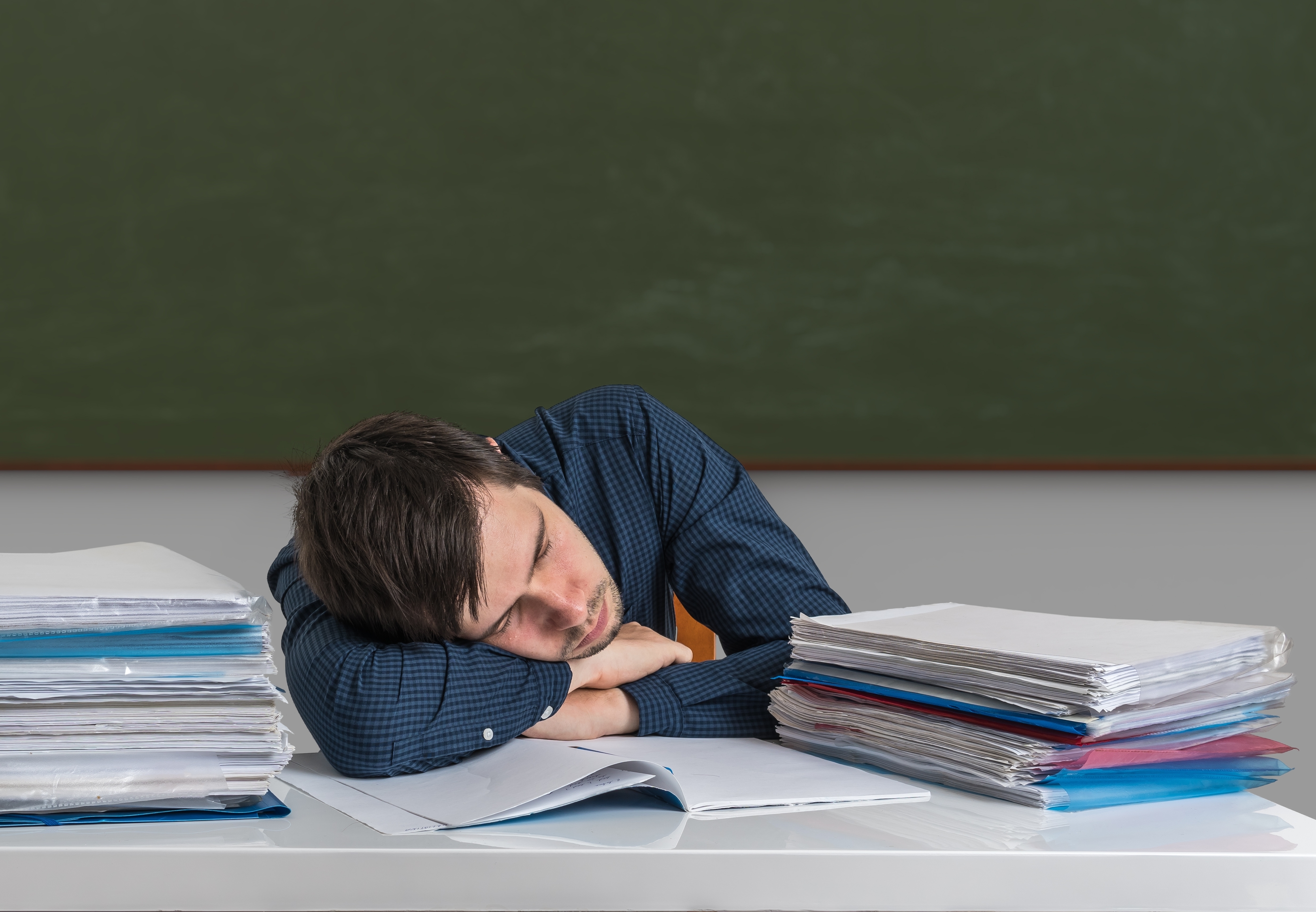 Man resting head on desk beside piles of paperwork, appearing exhausted or overwhelmed