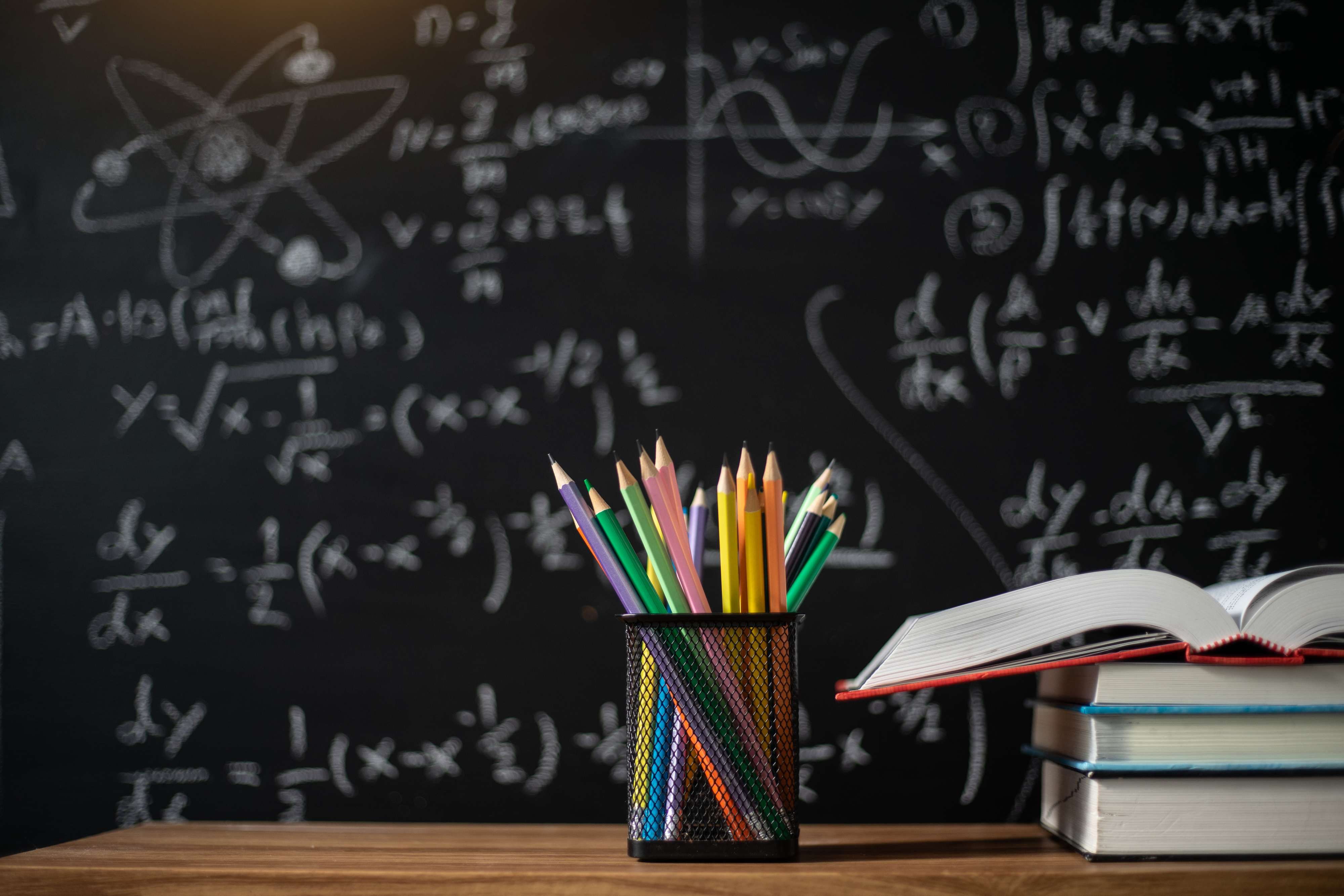 A variety of pencils in a holder with books on a table, in front of a blackboard with math equations