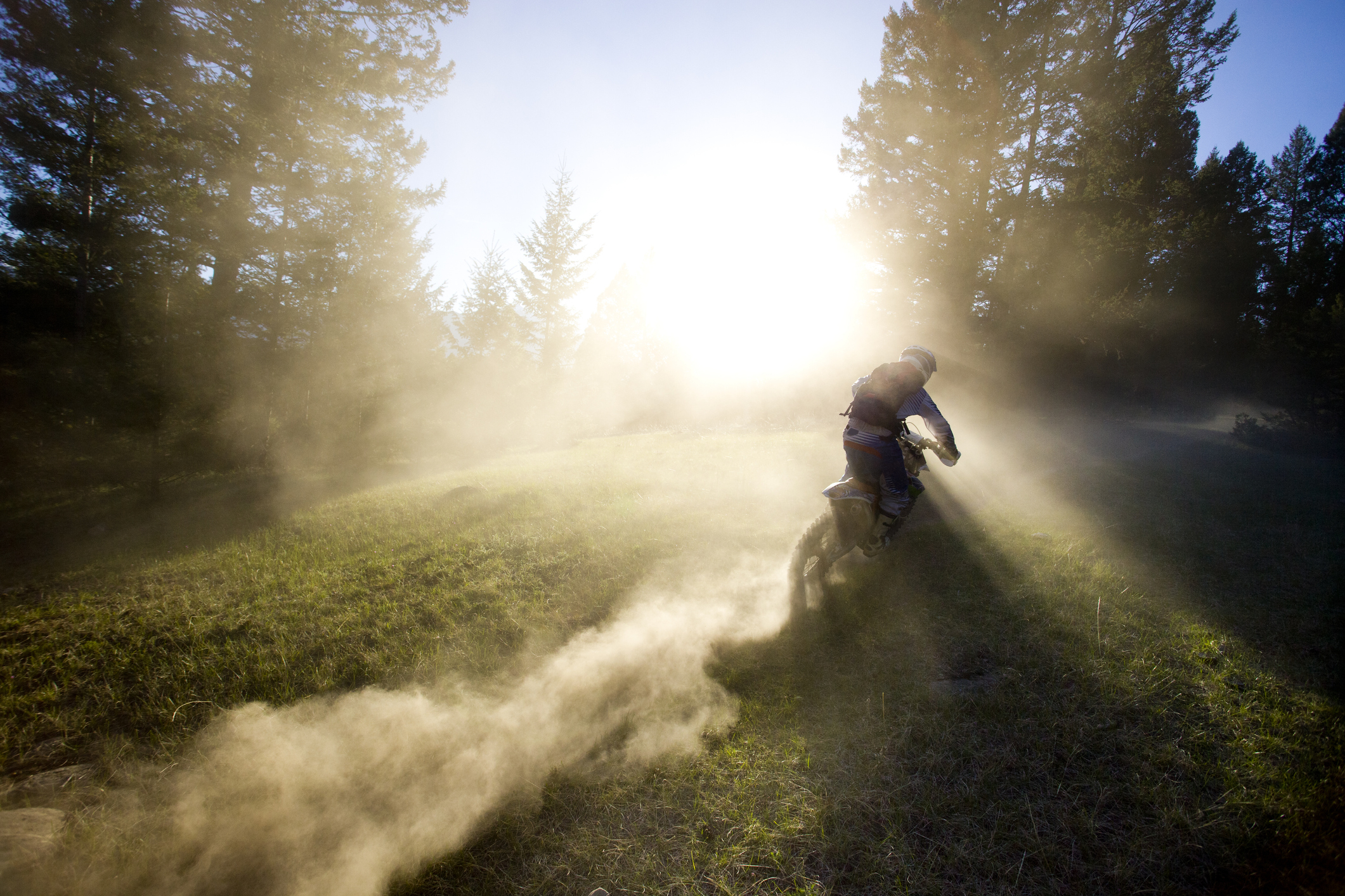 Person riding a dirt bike through a dusty trail with sunlight filtering through trees