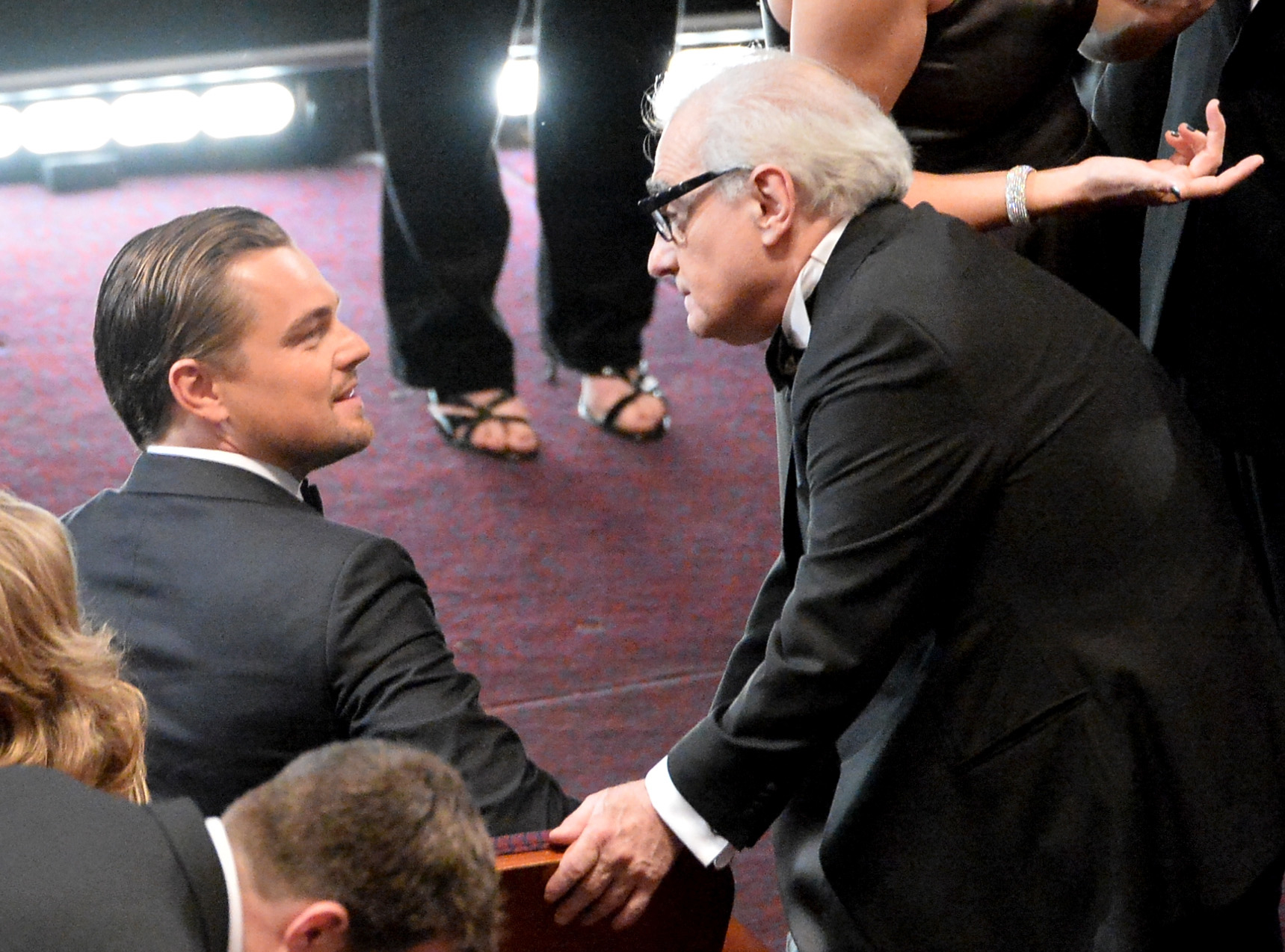 Leonardo DiCaprio seated, shaking hands with Martin Scorsese who is standing, both in formal suits at an event