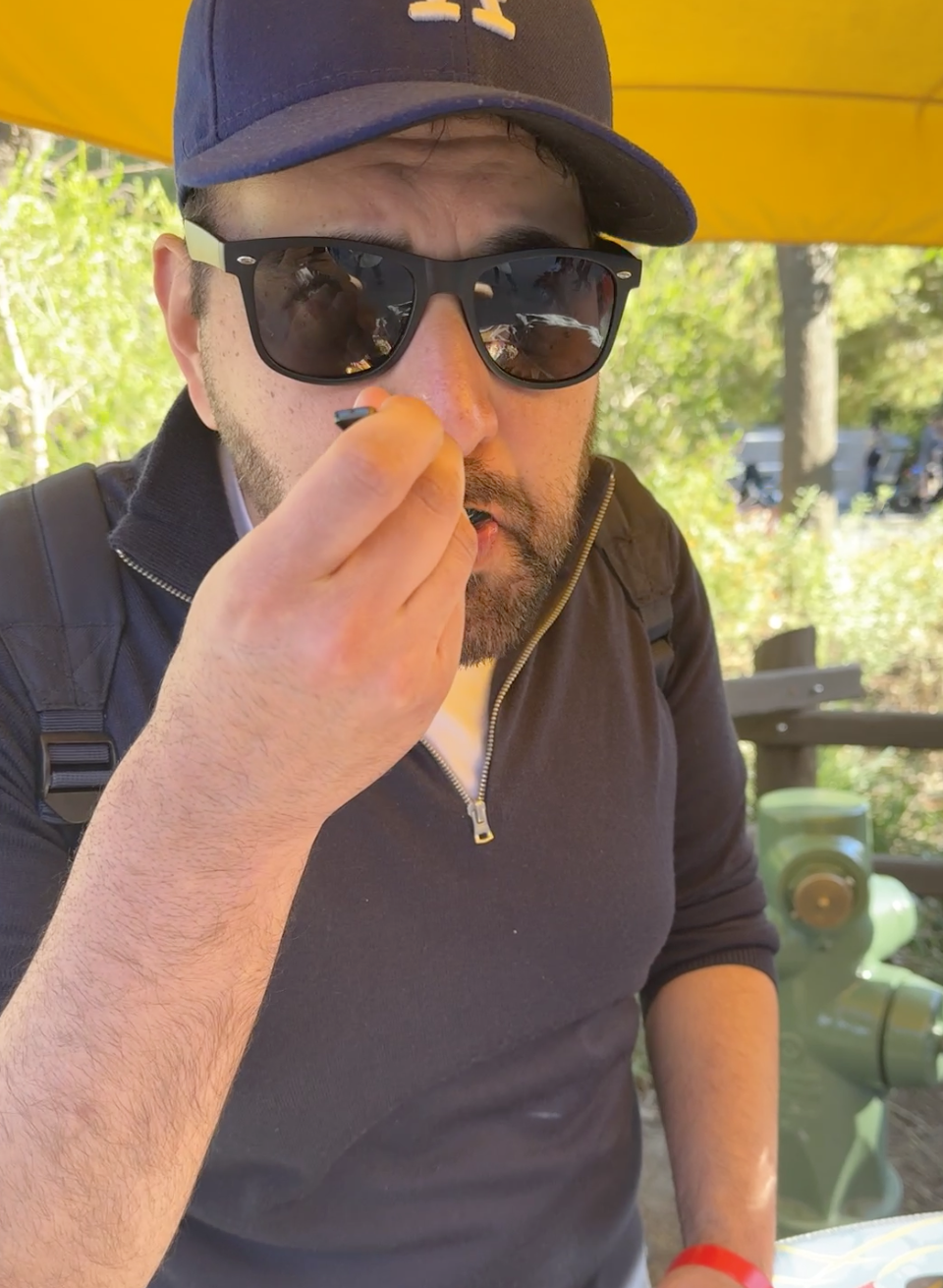 A man wearing sunglasses and a cap is eating under a yellow umbrella outdoors
