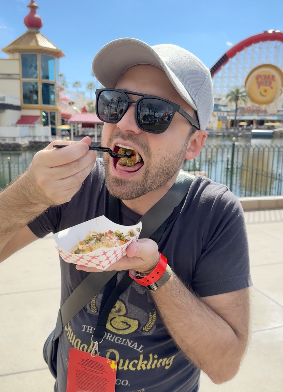 Person eating from a bowl at an outdoor amusement park, wearing a cap, sunglasses, and a graphic t-shirt