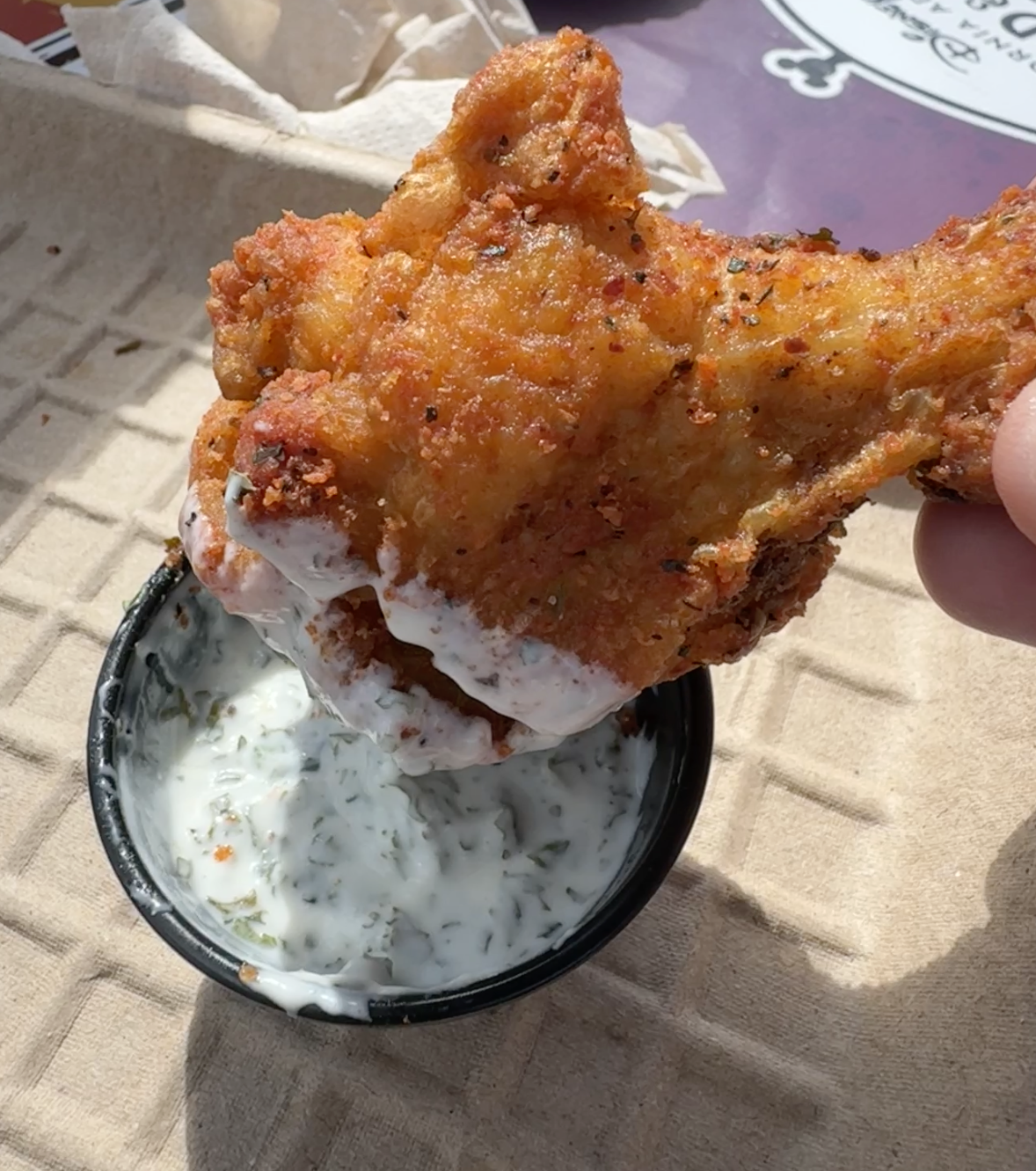 A hand dipping a piece of fried chicken into a cup of sauce, on a paper-lined plate
