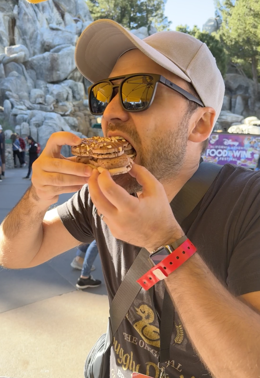 Man in sunglasses eats a large cookie at a theme park, surrounded by other visitors