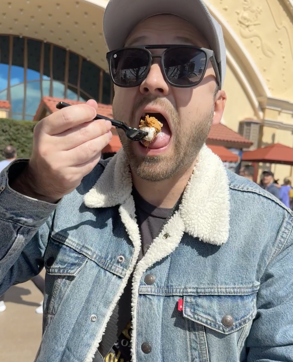 Man in sunglasses eating, wearing a jacket with a shearling collar, at an outdoor venue