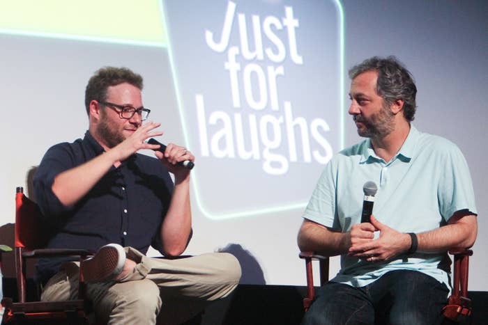 Two men on stage, one speaking into a microphone, in front of a &#x27;Just for Laughs&#x27; screen