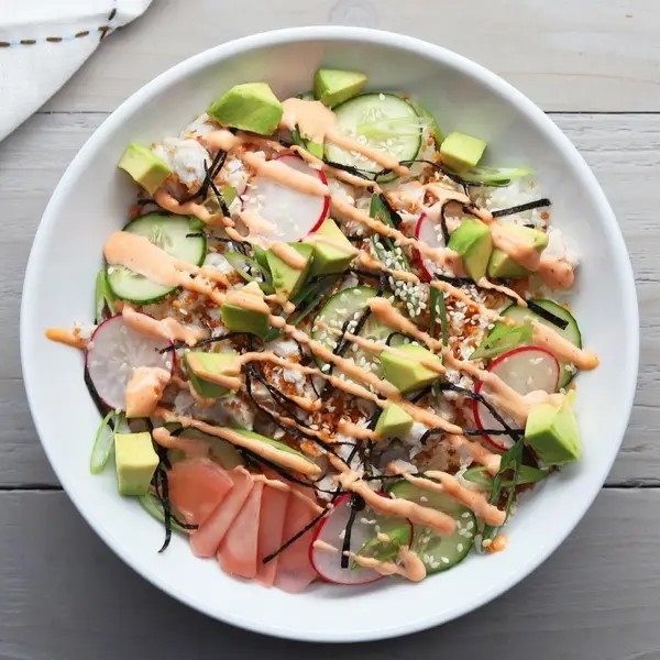 A sushi salad with avocado, cucumber, radish, sesame seeds, and drizzled sauce