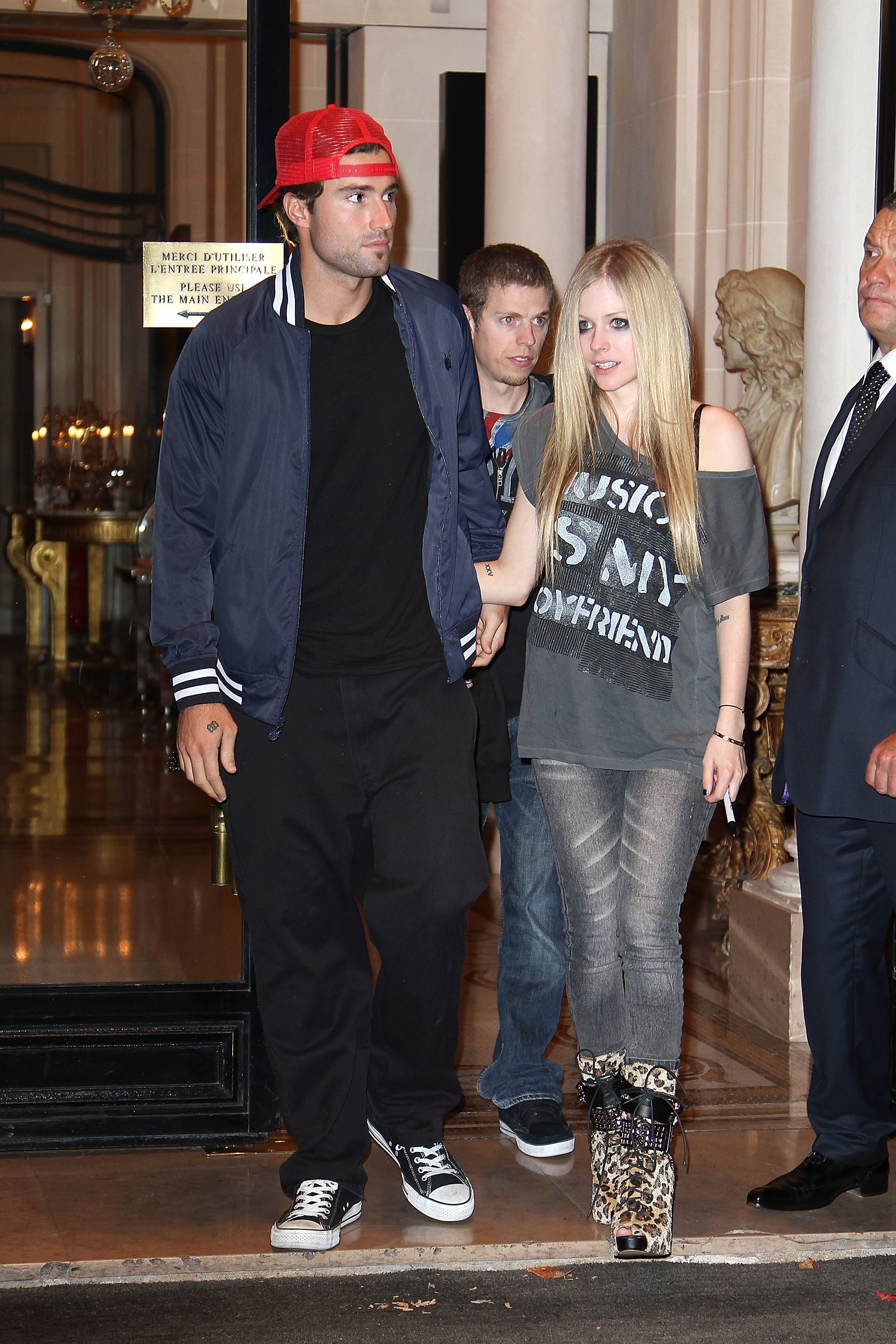 Brody in a cap and sneakers and Avril in a graphic tee and patterned pants exiting a building
