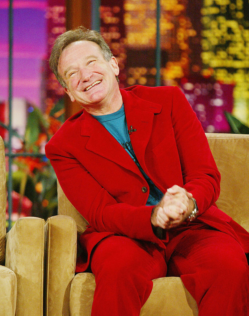 Robin Williams in a red suit sitting and laughing on a talk show set