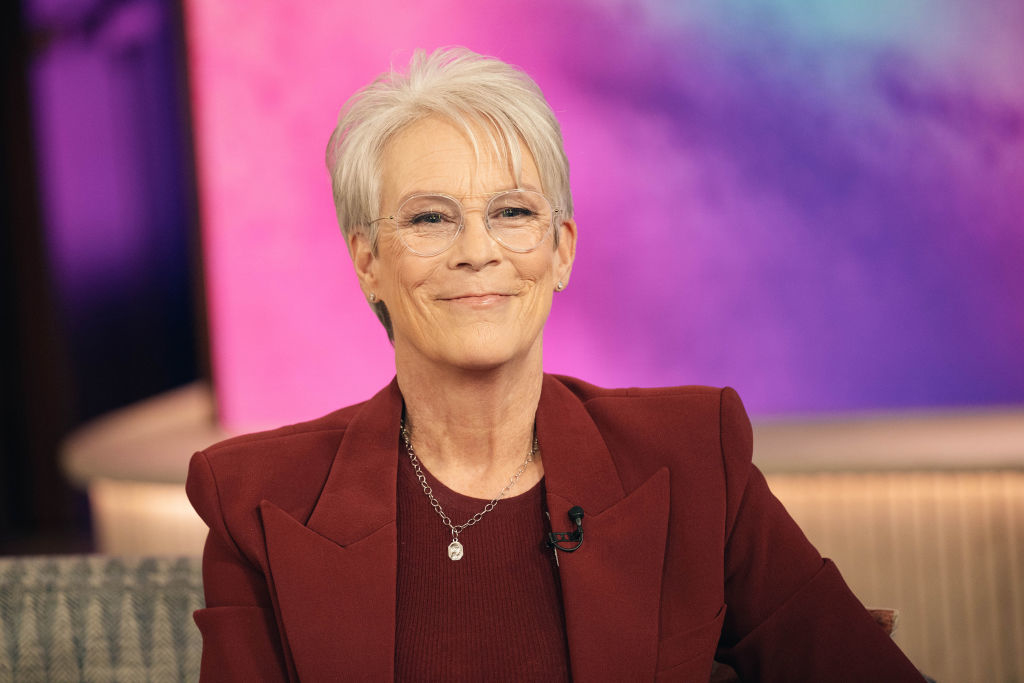 Jamie Lee Curtis sitting and smiling in a studio wearing a suit with a necklace