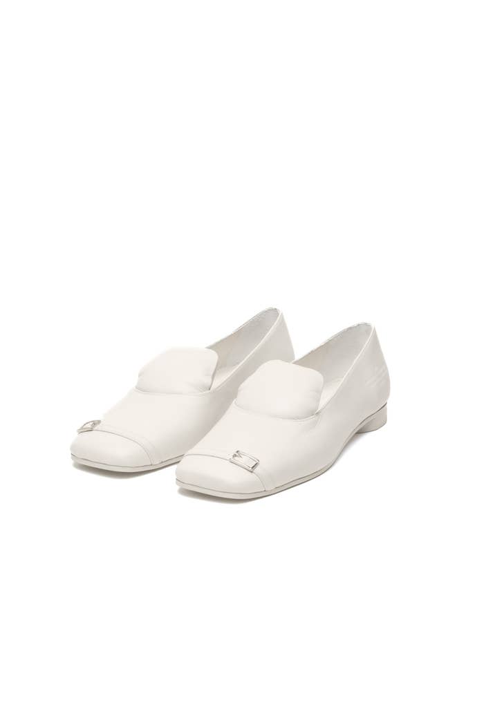 A pair of white loafers with a buckle detail on a neutral background