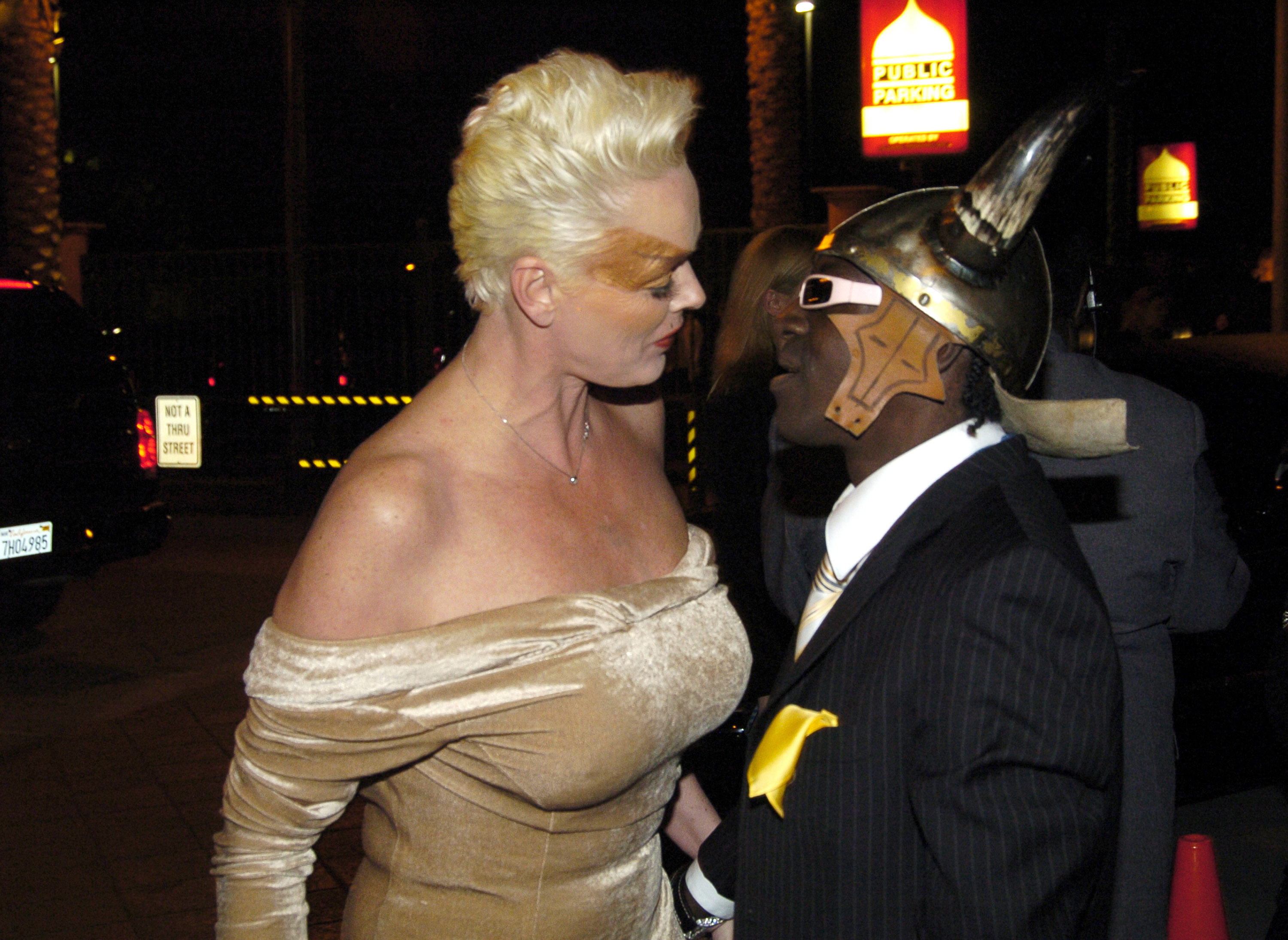 Brigitte in an off-shoulder dress looking at Flavor Flav, in a suit with a unique helmet