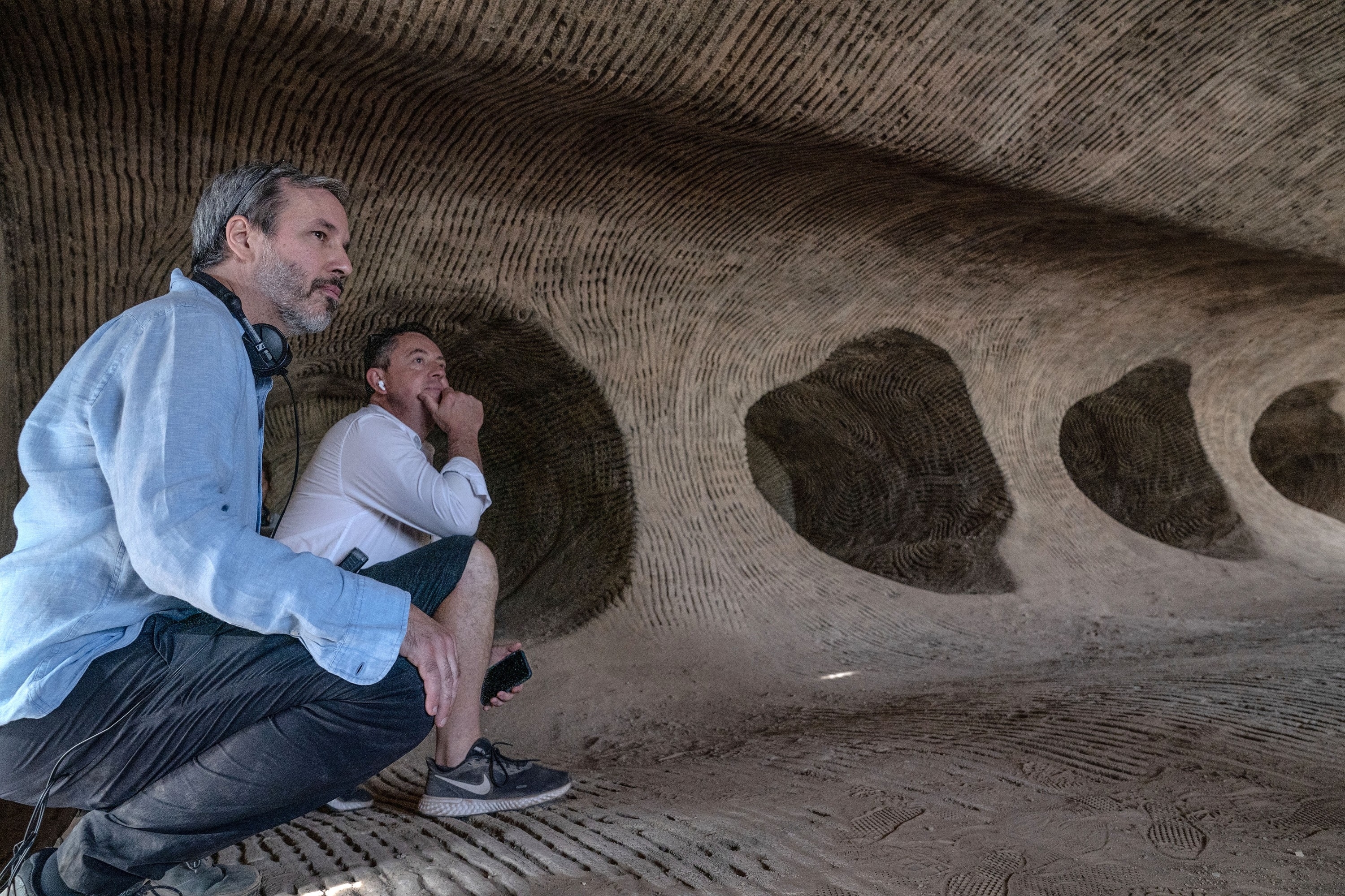 Denis Villeneuve crouching with another man inside a textured tunnel, looking thoughtful. They are on a film set