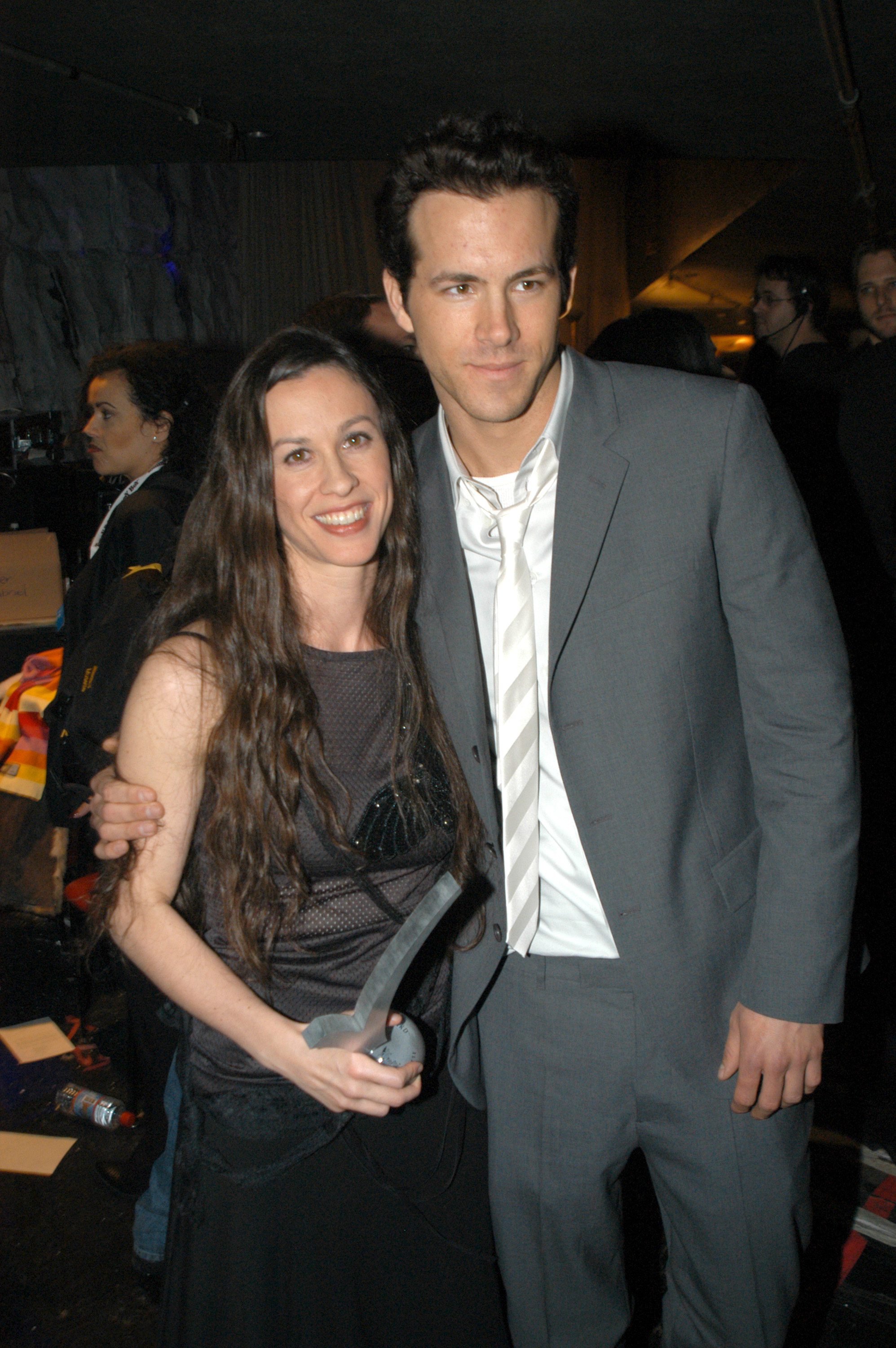 Alannis in a mesh top and holding an award, and Ryan in a suit with an unbuttoned shirt, with his arm around her