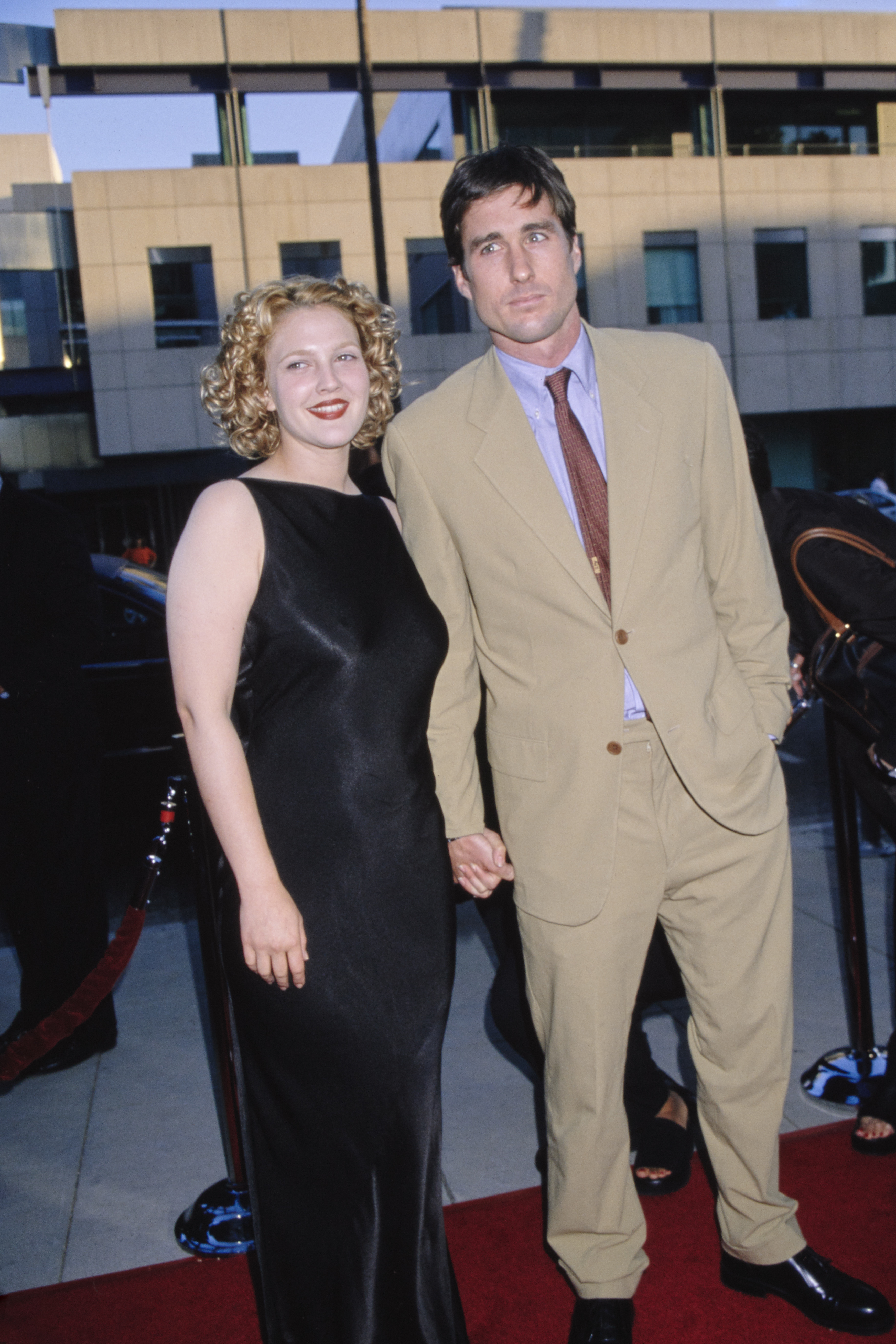 Drew with curly hair wearing a sleeveless black gown, and Luke in a beige suit and tie, holding hands on the red carpet