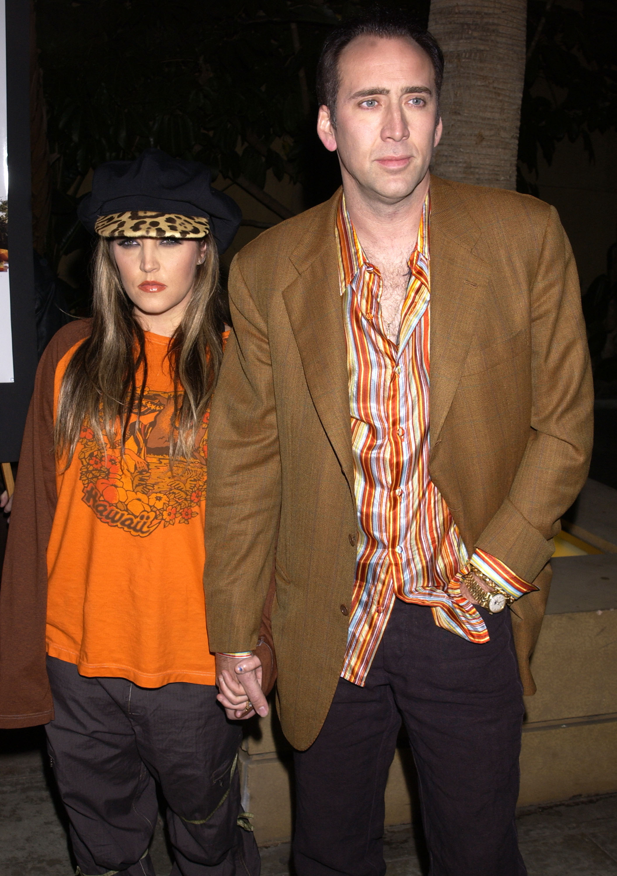 Lisa Marie in a printed shirt and blazer, and Nic in a cap and layered shirts, holding hands