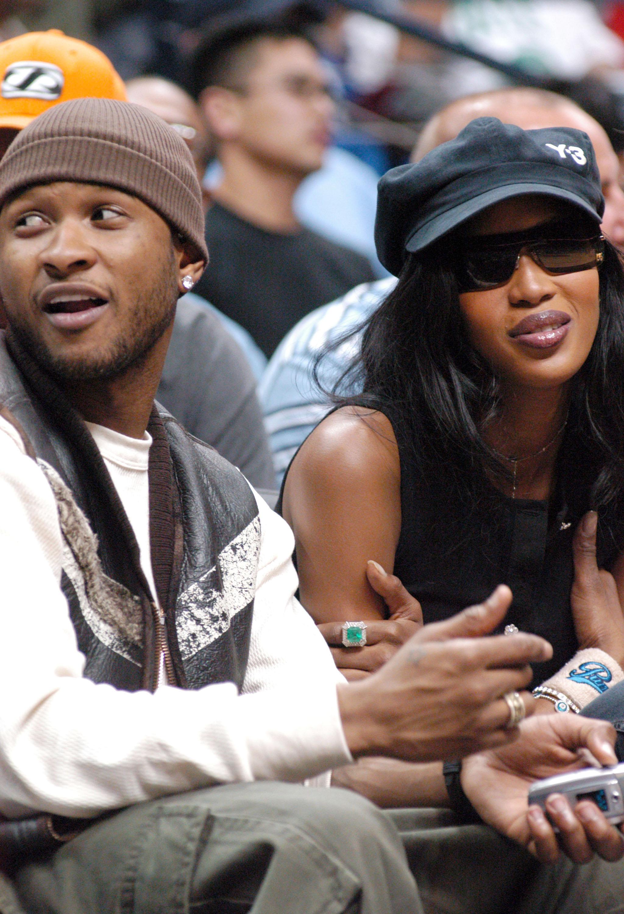 Usher wearing a graphic shirt and beanie, and Naomi in a black cap and sleeveless top, sitting at a sporting event