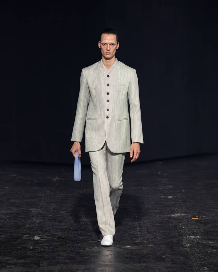 Model on runway in fitted light-toned suit with button-up jacket, straight-leg trousers, and white shoes