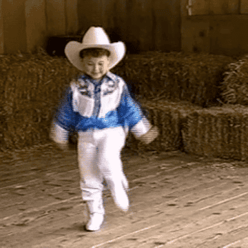 Niño con traje de vaquero con sombrero, chaqueta con flecos y botas, caminando en un establo
