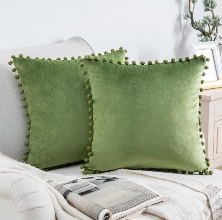 Two green square pillows with pom-pom edges on a couch, next to a magazine