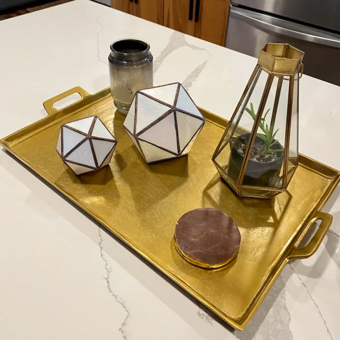 A decorative tray on a counter with two geometric candle holders, a plant, a jar, and coaster
