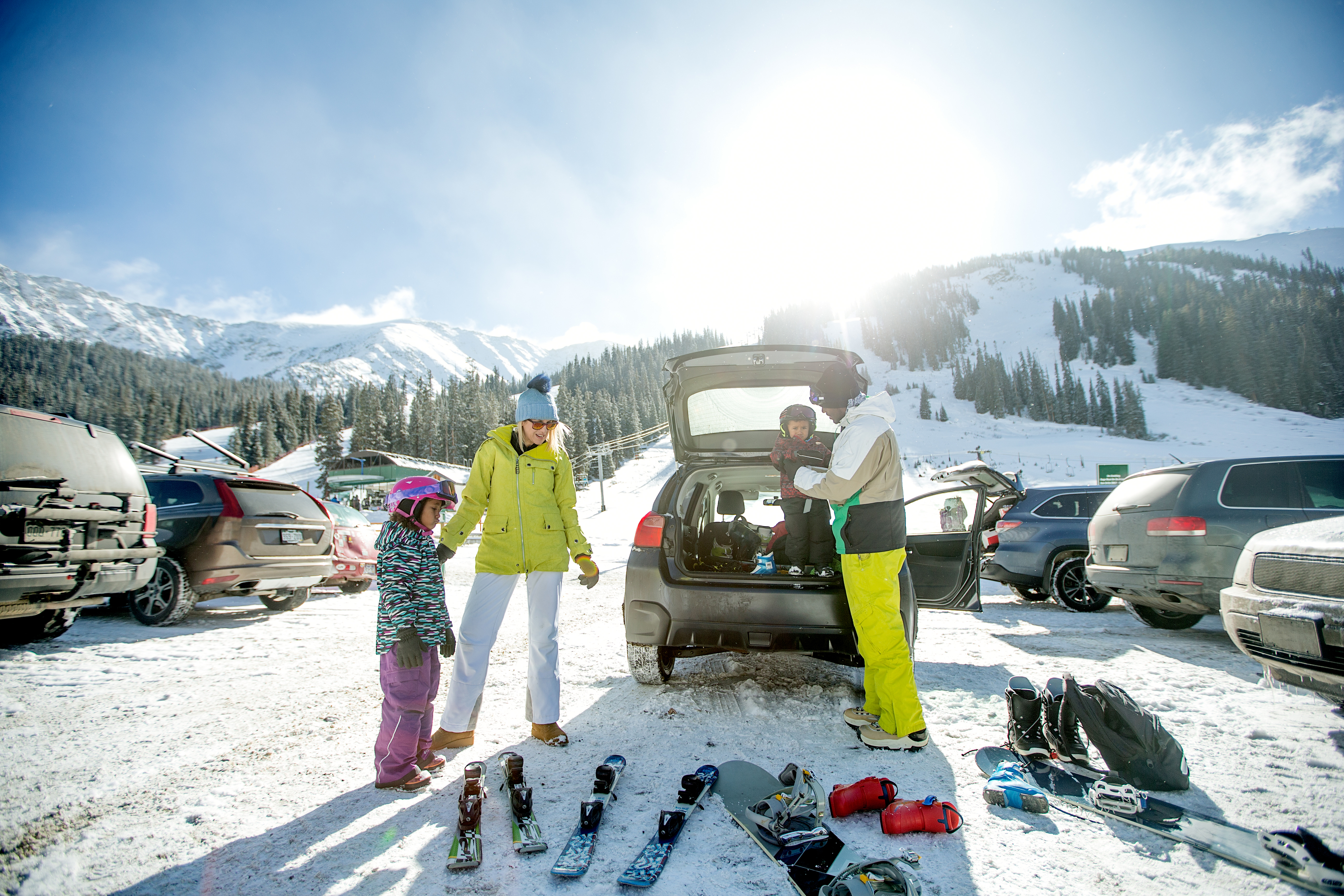 Three people in ski gear by a car in a snowy parking lot, preparing for skiing