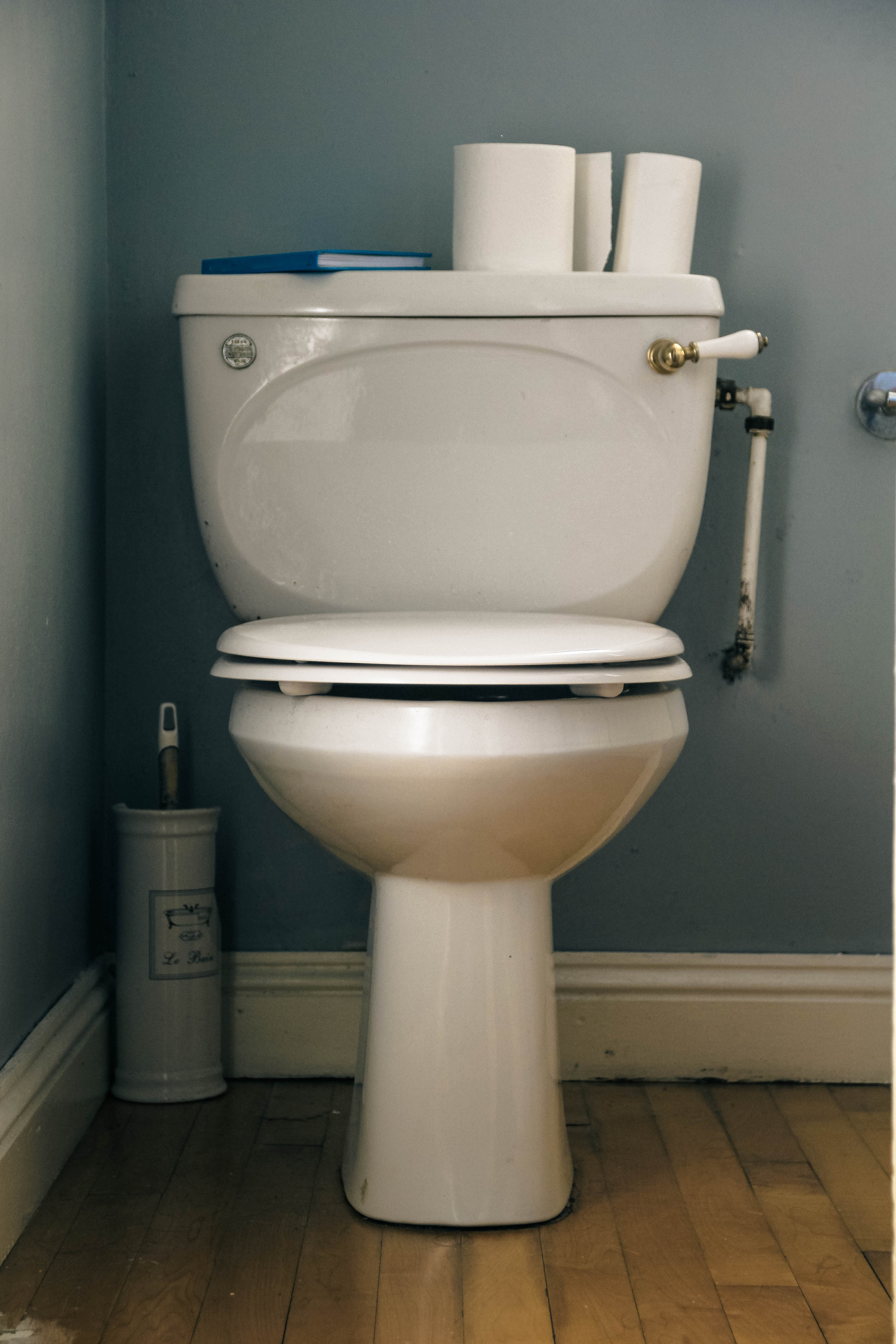 Toilet with a closed lid and extra rolls of paper on the tank, next to a small trash can