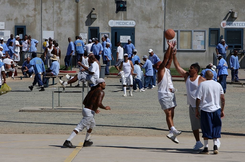 Inmates playing basketball in a prison yard with others watching