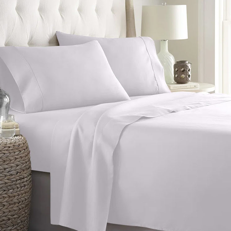 A neatly made bed with white bedding and pillows, a lamp, and decor on the bedside table