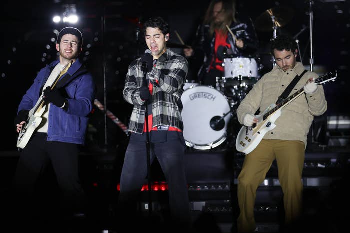 Jonas Brothers performing on stage with microphones and guitars, casual attire. Drummer in background