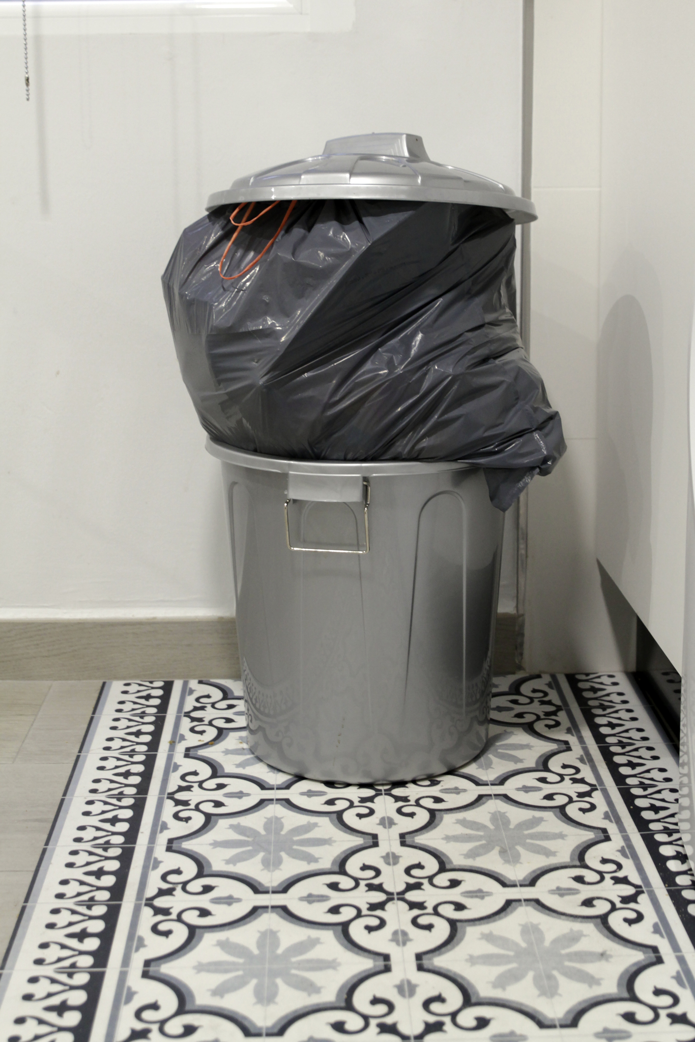 A full trash can with a closed lid and a black bag sticking out in a kitchen