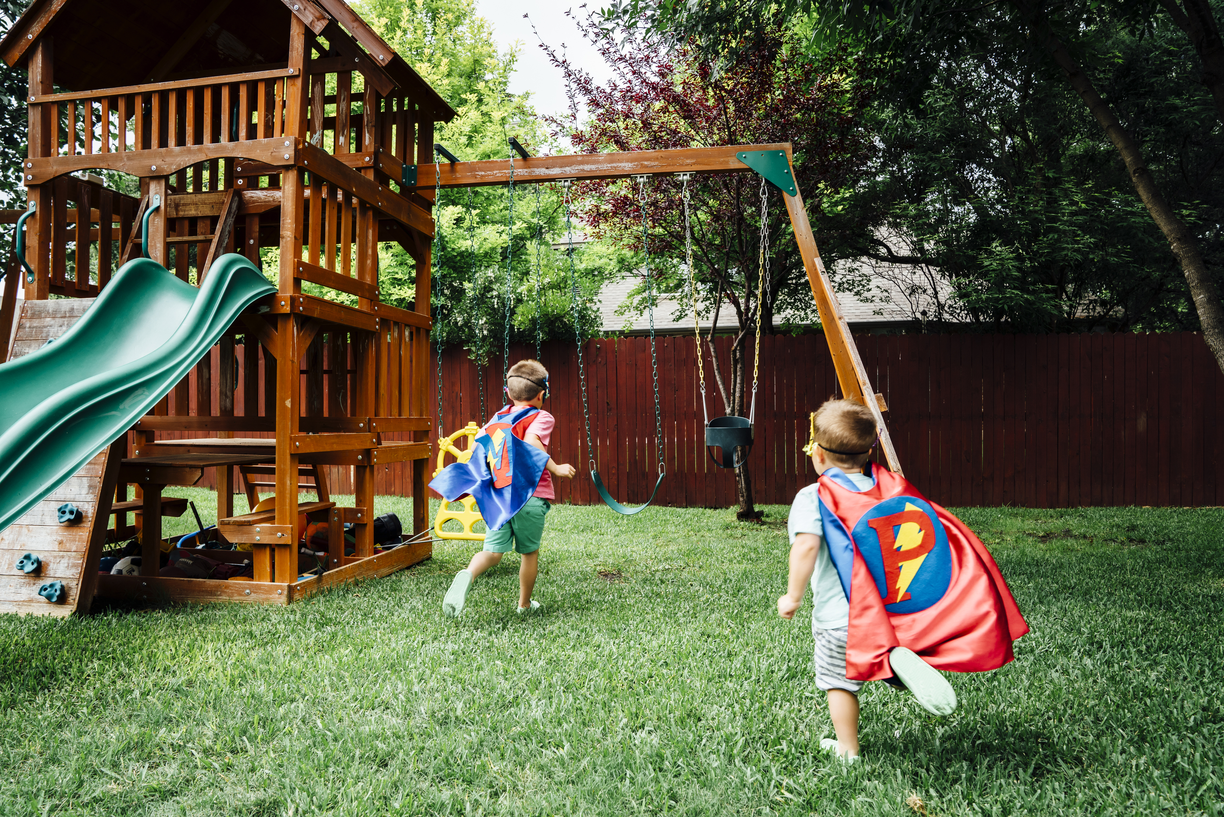 Two children wearing superhero capes play near a swing set in a backyard