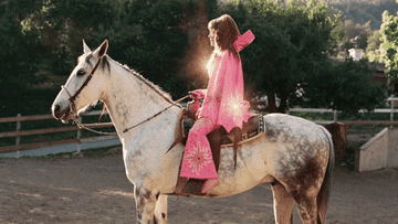 Mujer monta a caballo, luce un poncho rosa con adornos; ambiente rural al atardecer