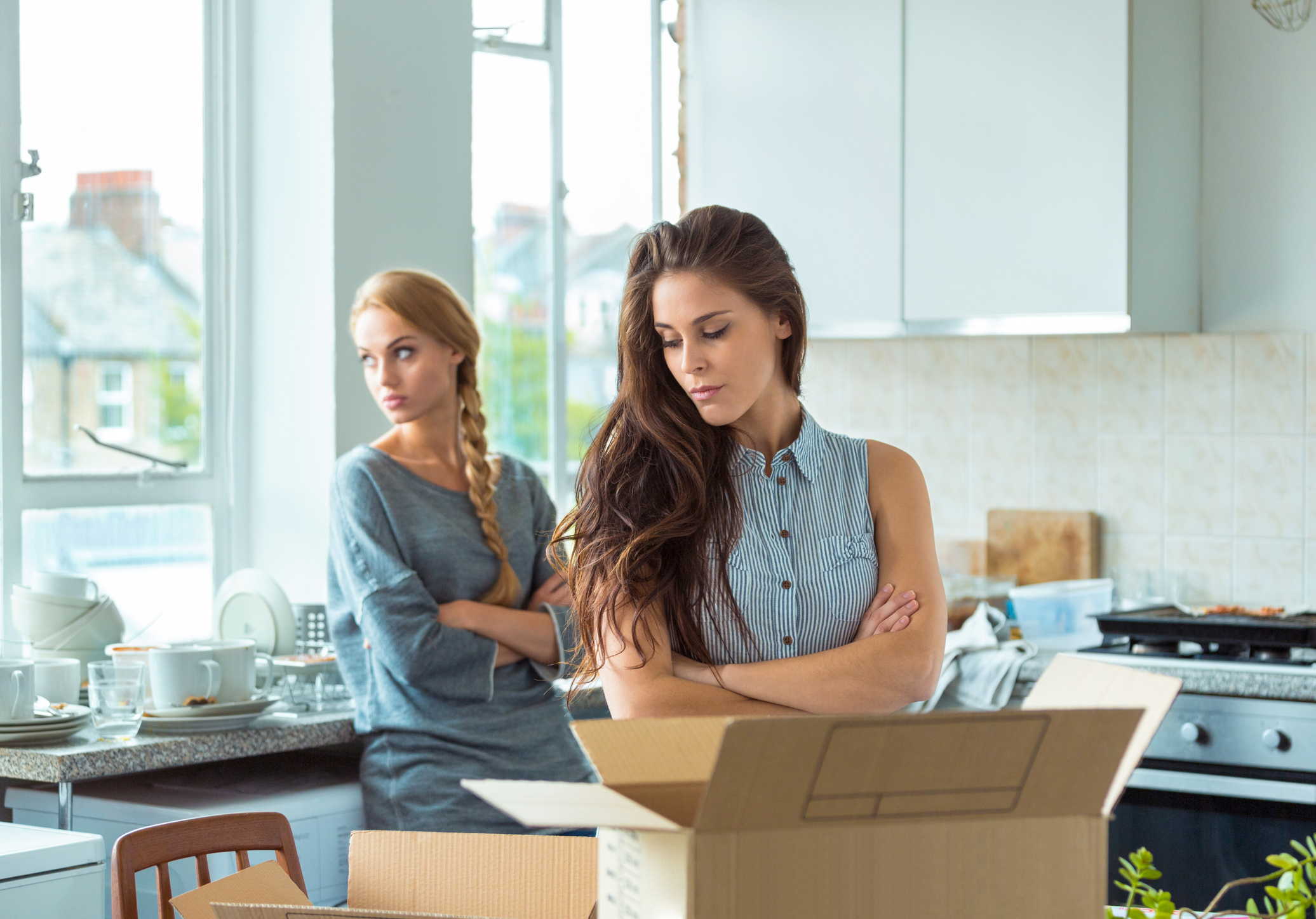 Two women in a kitchen, one standing with arms crossed looking at the other who is focused on an open cardboard box