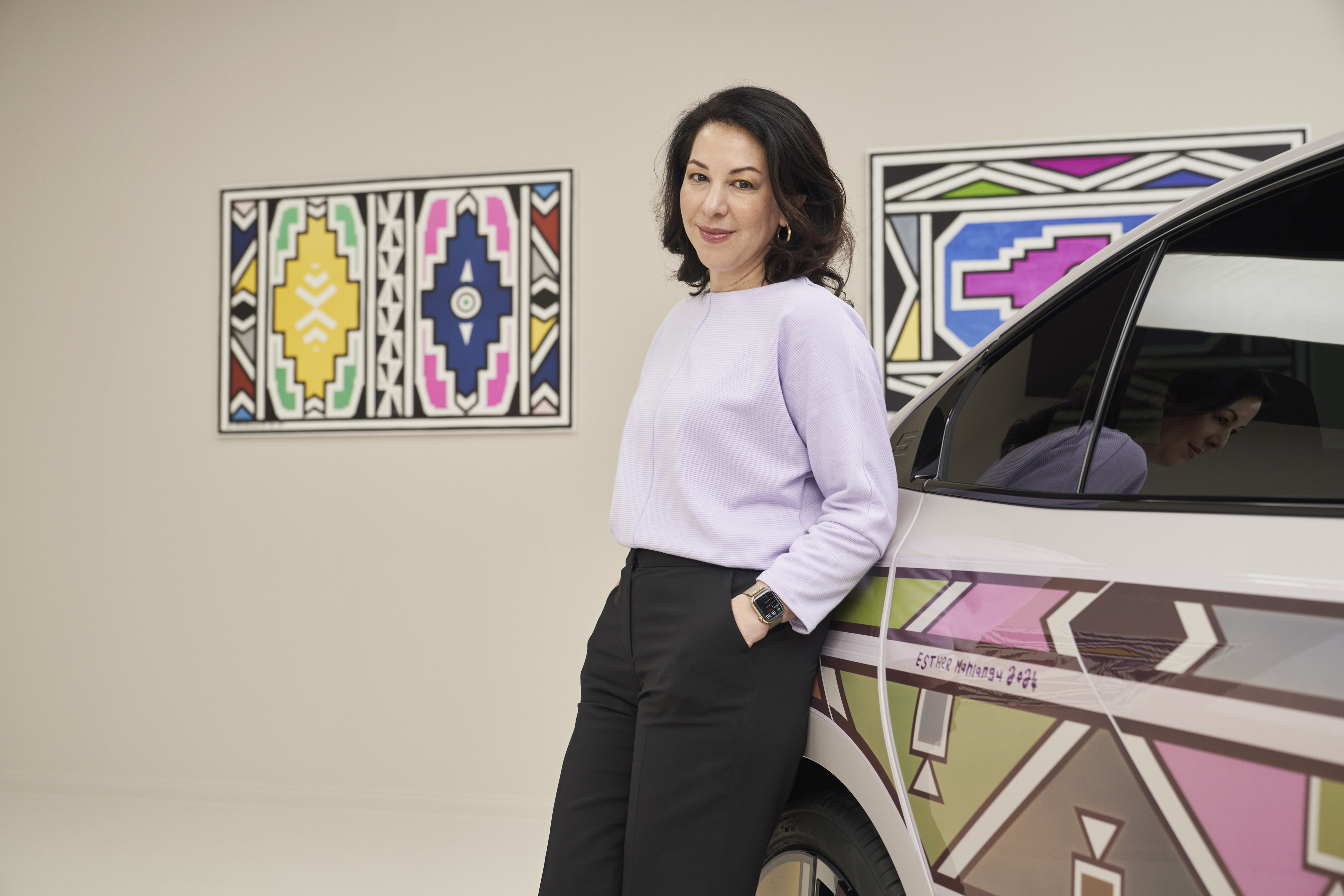 Woman in a purple top and black pants standing beside a car with graphic patterns, artwork on the wall behind