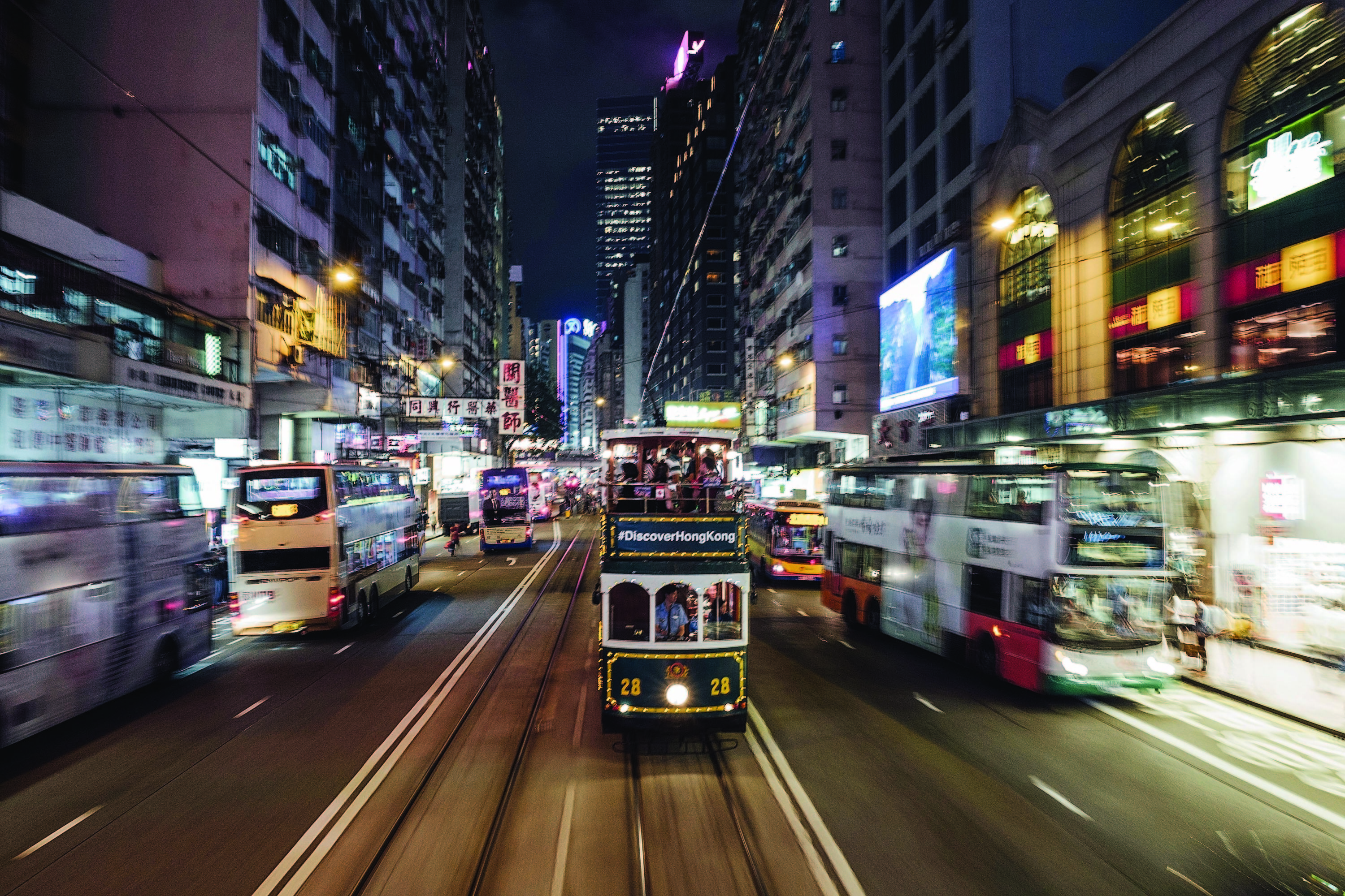City street scene with a tram and buses, illuminated advertisements on buildings, and a #DiscoverHongKong banner