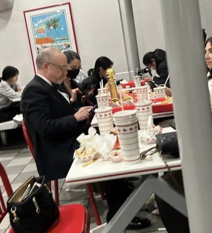 Paul Giamatti sitting at a fast food restaurant with a table full of food containers, dressed in a suit, holding a smartphone