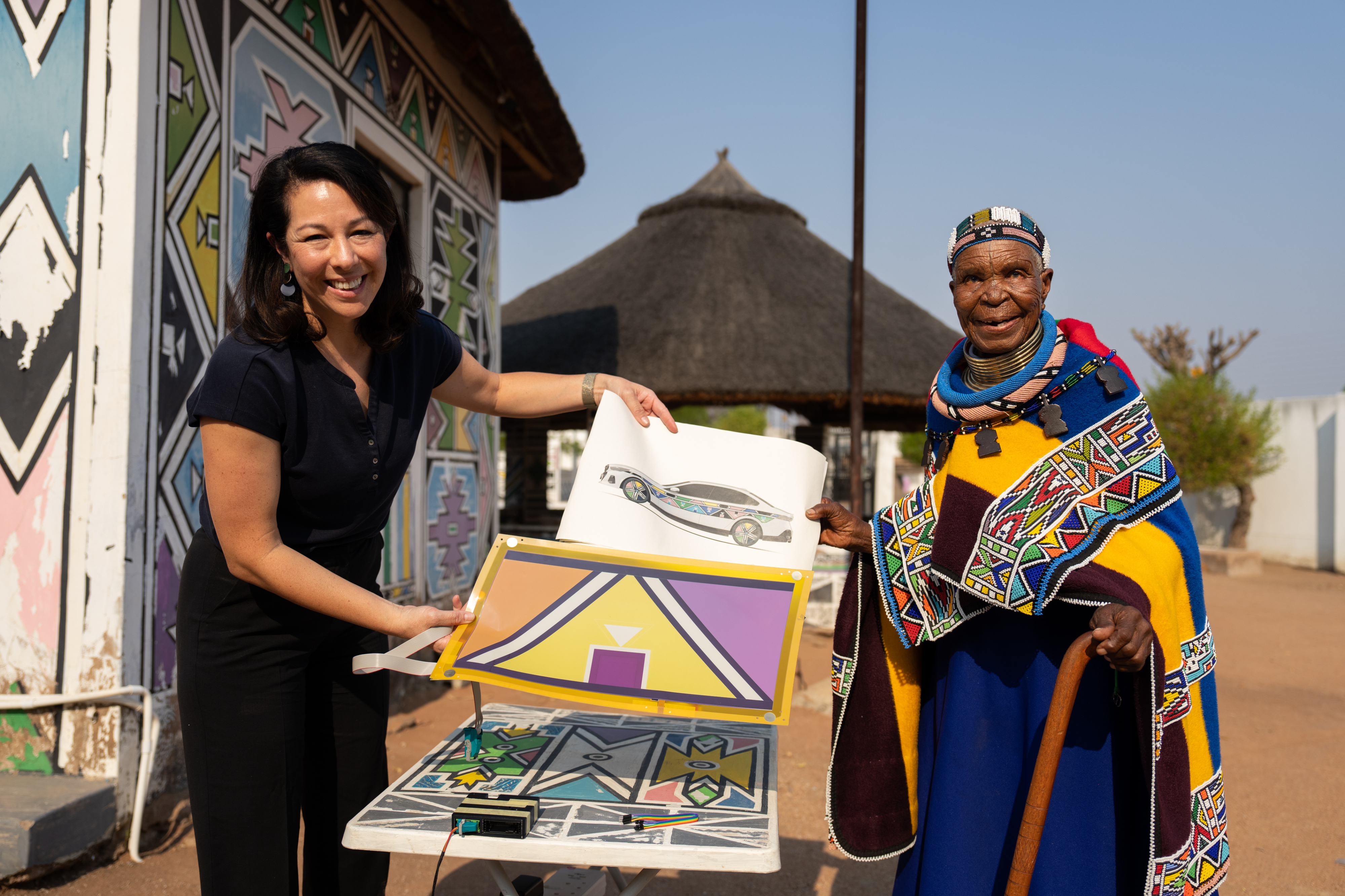 Two individuals presenting a piece of artwork outside with traditional hut and mural in the background. One is wearing cultural attire