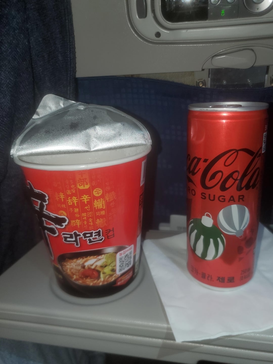 Swollen instant noodle cup beside a Coke Zero can on an airplane tray table