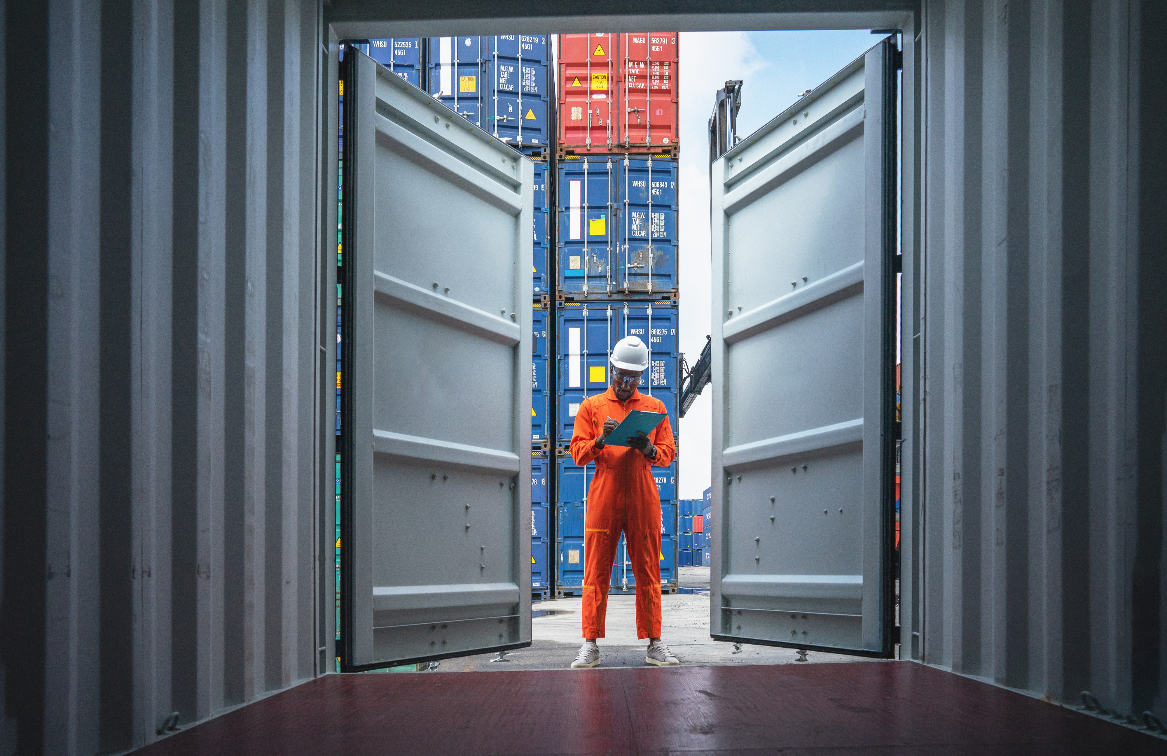 Worker in safety gear inspecting clipboard inside open cargo container with stacked containers in background