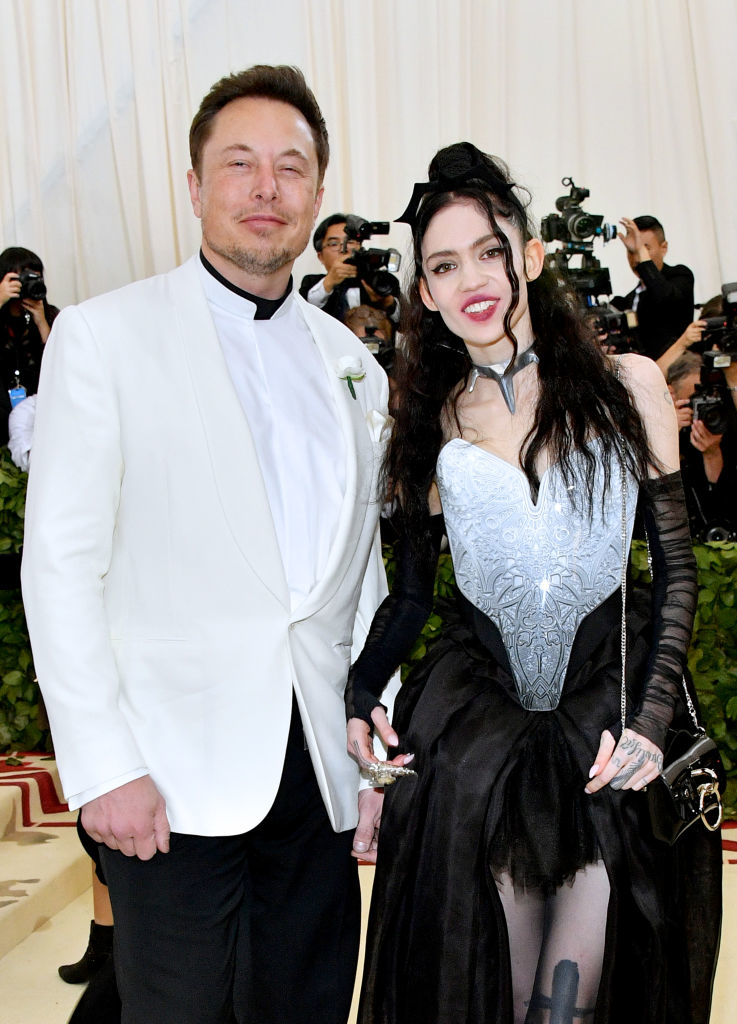 Elon Musk in a white tuxedo with a flower pin, standing beside Grimes in a black and silver outfit with unique details. They are at an event