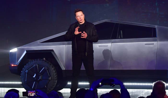 Elon Musk speaking at Tesla Cybertruck unveiling. He wears a black leather jacket and black shirt. Truck in the background, audience in the foreground