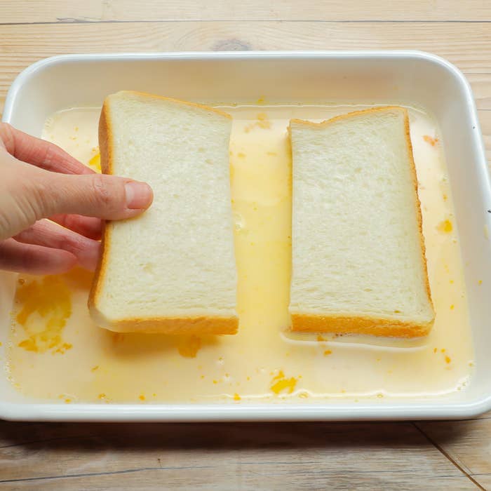 手がフレンチトーストの生地に浸したパンを浸けている様子。
