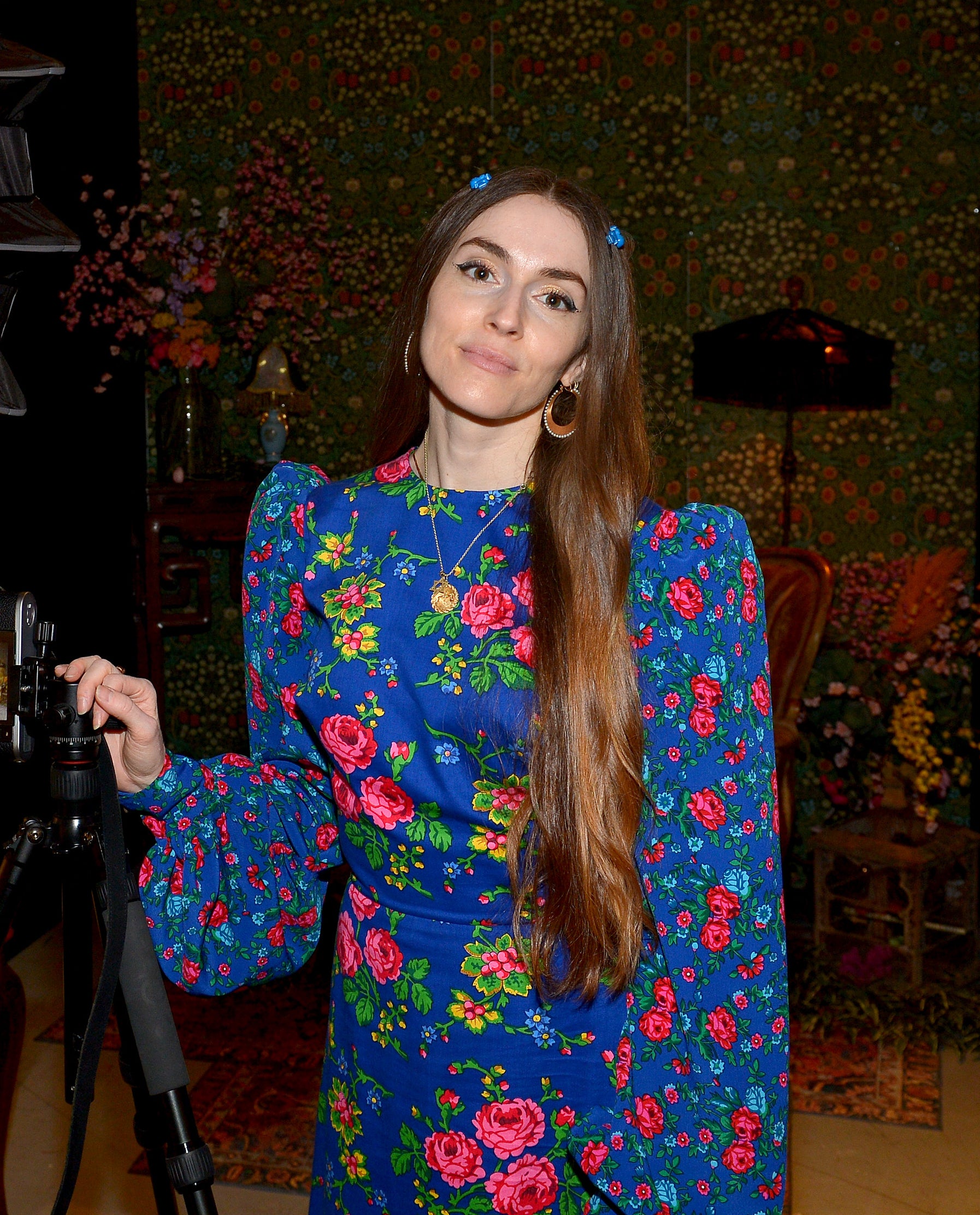 Anna Marie in a floral dress stands by a camera and lighting equipment, slightly smiling