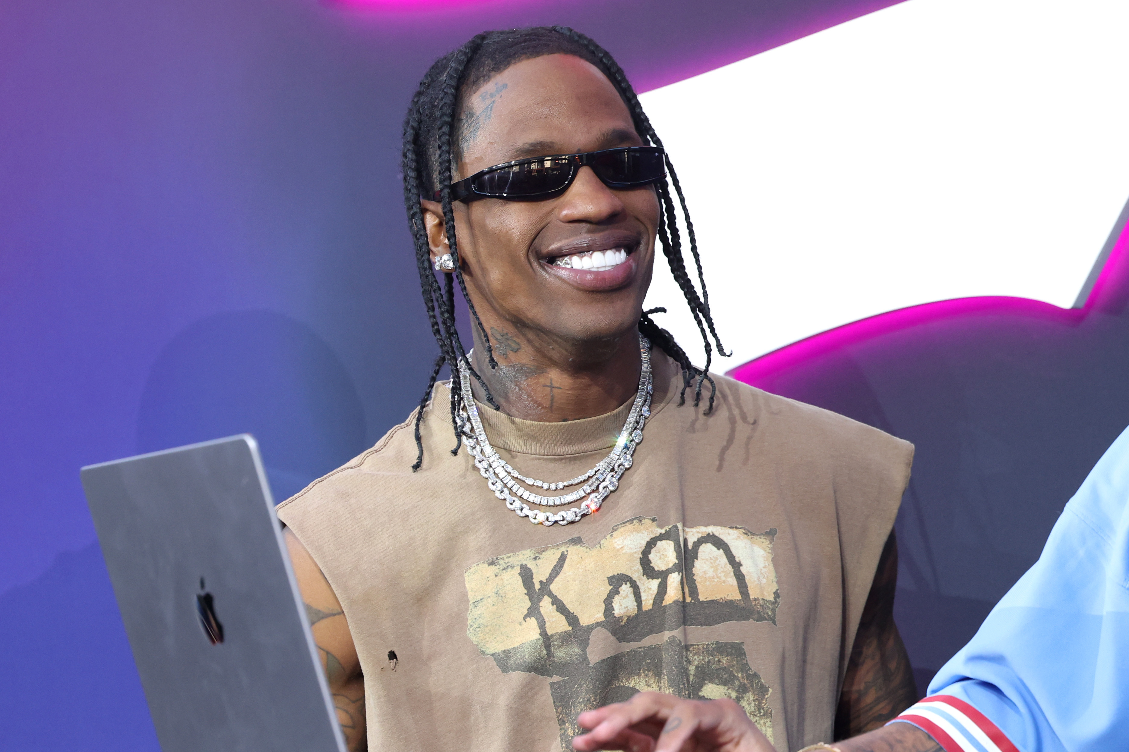 Travis Scott smiling, wearing a graphic tee and layered necklaces, stands next to a man with a laptop