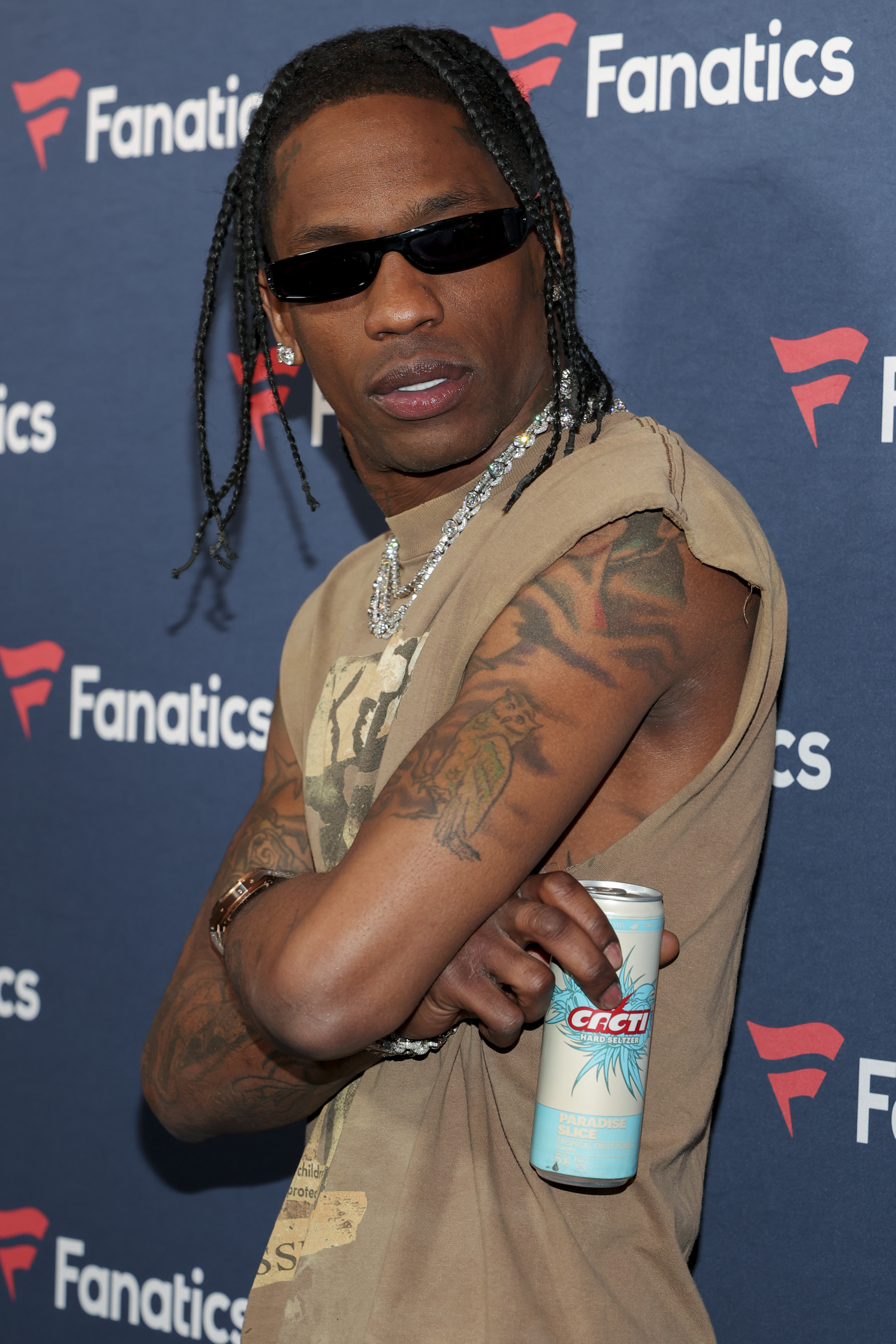 Travis Scott in a sleeveless top and chains, holding a can, with tattoos visible, posing
